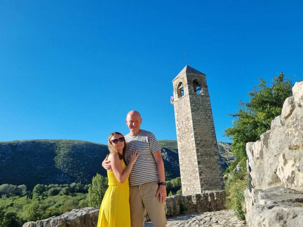 Para turystów na tle kamiennej wieży zegarowej Sahat-kula w miasteczku Počitelj na wzgórzu.