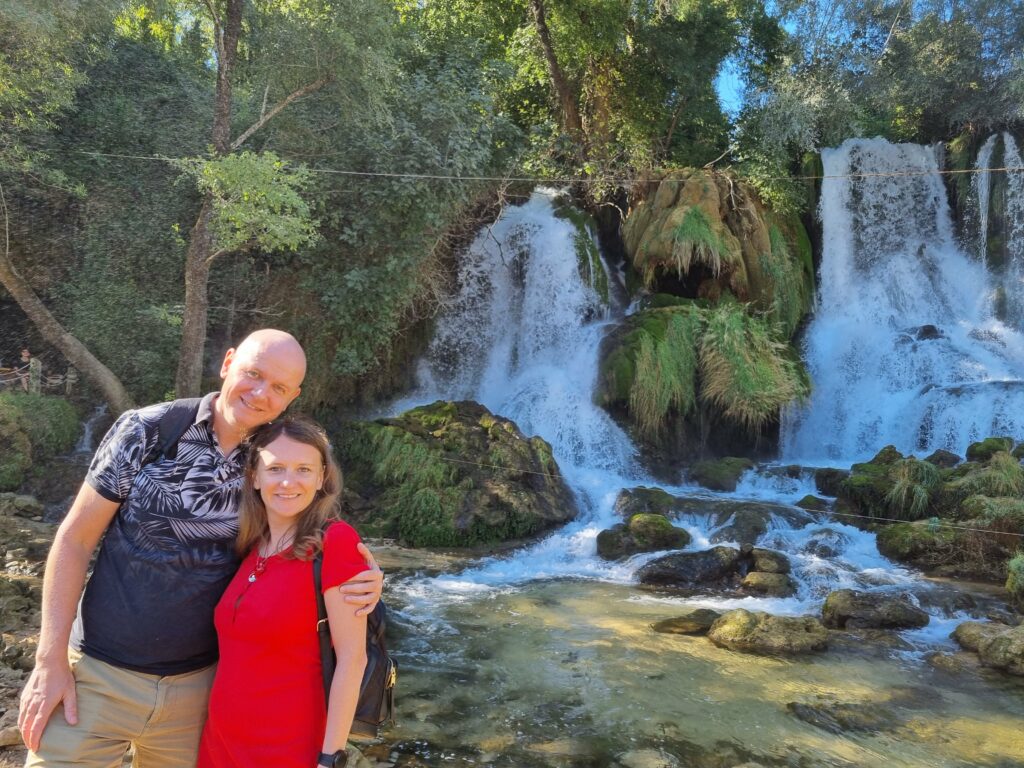 Para turystów stojąca na drewnianym pomoście przed potężnymi kaskadami Wodospadów Kravica w BiH.