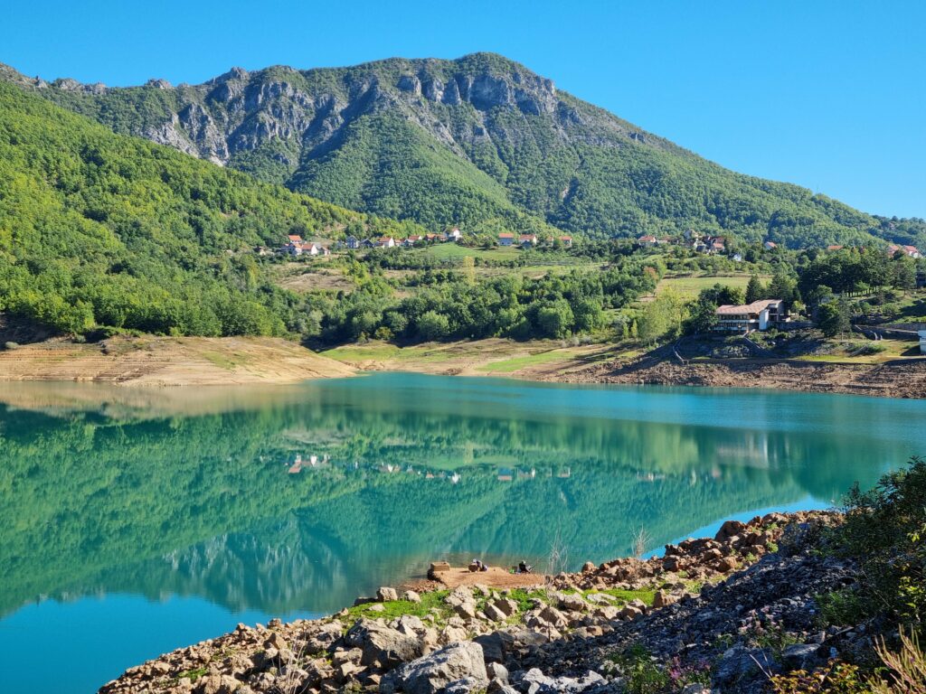 Zbliżenie na brzeg Jeziora Ramsko z widoczną piaszczystą linią brzegową i turkusową, czystą wodą w słoneczny dzień.
