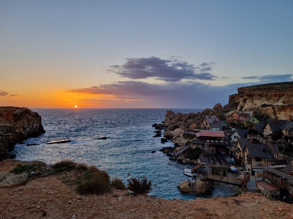 Zachód słońca nad Anchor Bay na Malcie, z widokiem na klify i kolorowe zabudowania Popeye Village.