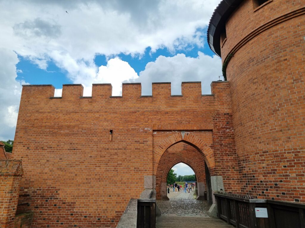 Ostrołukowa brama przejazdowa w ceglanym murze z krenelażem na Zamku w Malborku, w tle spacerujący turyści.