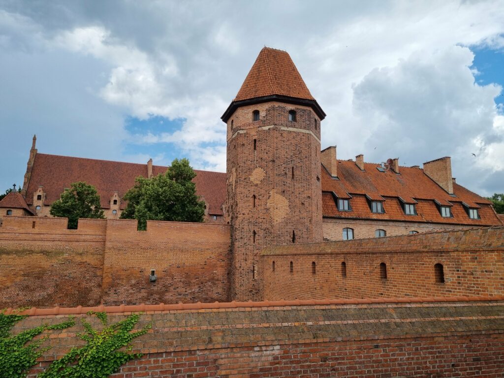 Ceglane mury obronne i masywna baszta z czerwoną dachówką na terenie Zamku Krzyżackiego w Malborku.