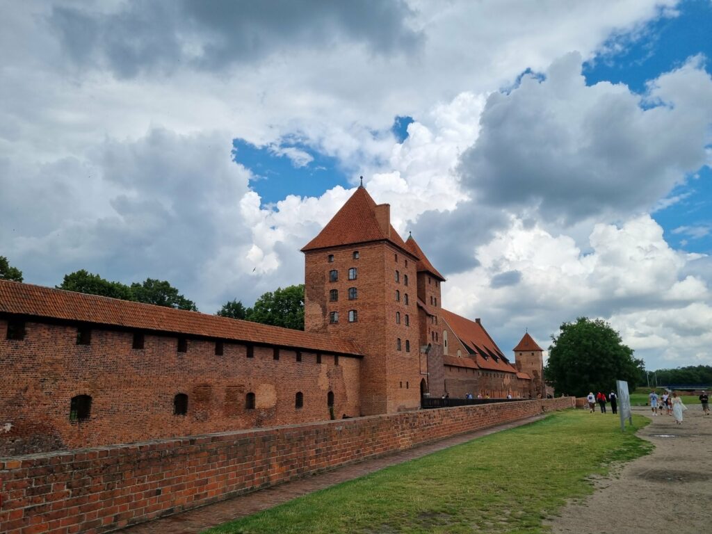 Długie, ceglane mury zewnętrzne i wysokie wieże Zamku w Malborku widziane z perspektywy ścieżki spacerowej.
