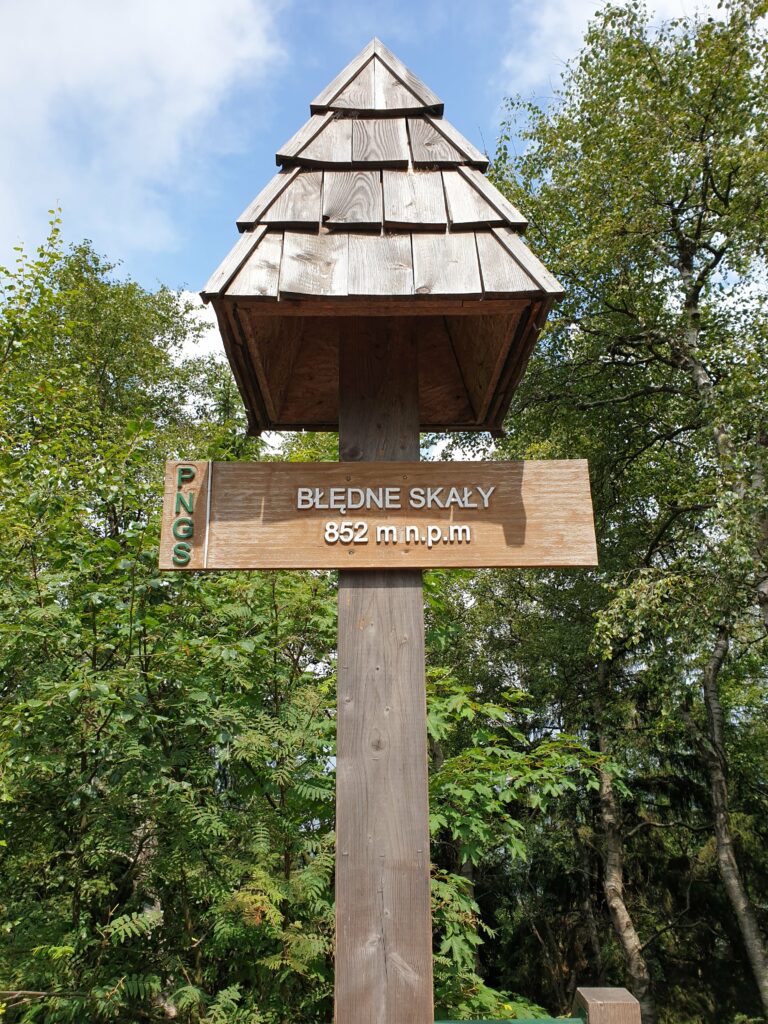 Drewniana tabliczka informacyjna z napisem Błędne Skały 852 m n.p.m. na tle lasu w Górach Stołowych.