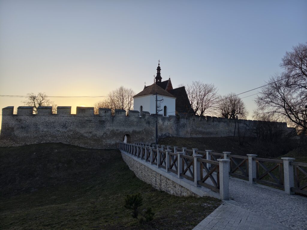 Drewniany most przerzucony nad dawną suchą fosą, prowadzący do kamiennego wejścia w średniowiecznych murach obronnych obok kościoła w Szydłowie.