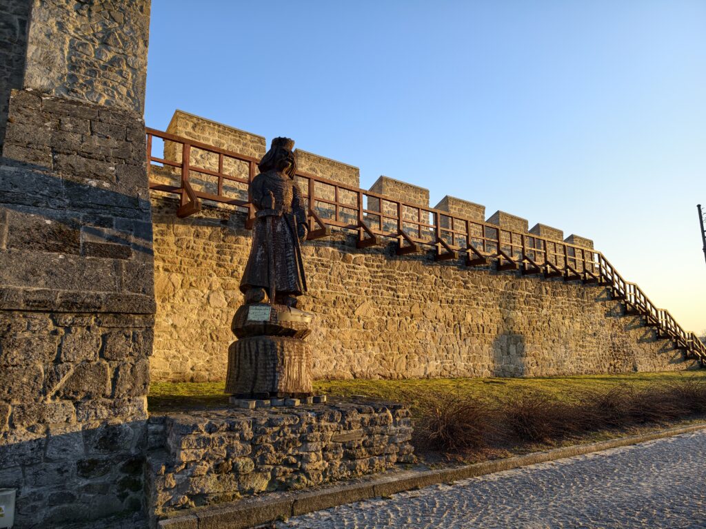 Wysokie, kamienne mury obronne w Szydłowie z widocznymi blankami i basztami na tle błękitnego nieba.