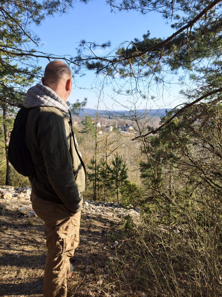 Turysta stojący na zalesionym odcinku szlaku, podziwiający rozległą panoramę Gór Świętokrzyskich widoczną przez prześwity między drzewami.