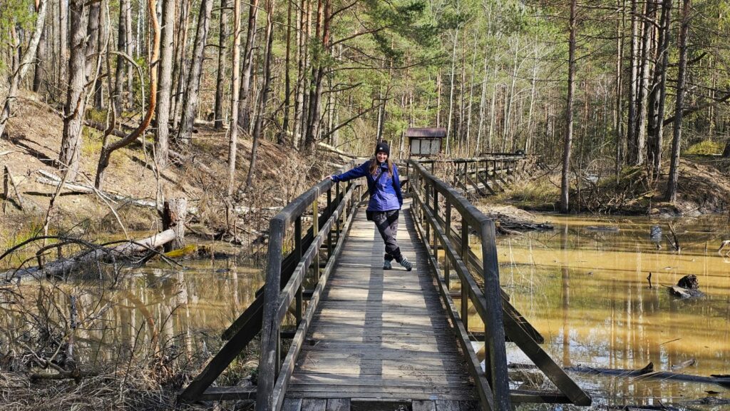 Turystka spacerująca po drewnianej kładce w rezerwacie Gagaty Sołtykowskie, w otoczeniu lasu i charakterystycznej rdzawej ziemi.