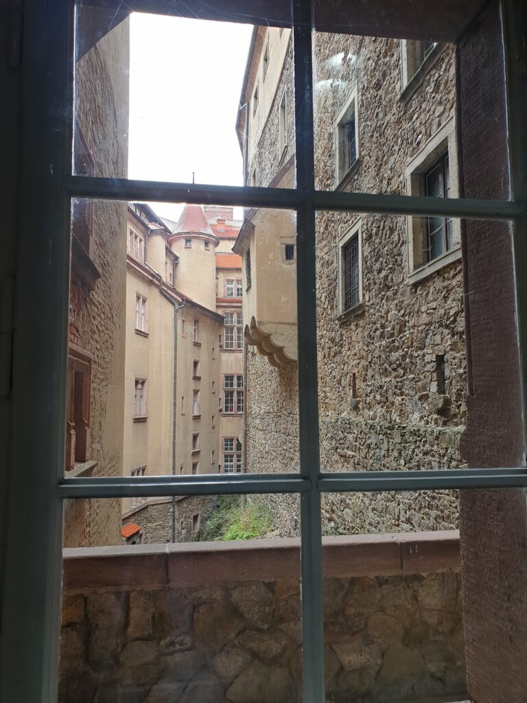 Widok przez okno na wewnętrzne mury, wieżyczki i strome dachy Zamku Książ w Wałbrzychu. Kadr ukazuje różnorodne style architektoniczne i wielowiekową historię rozbudowy kompleksu.