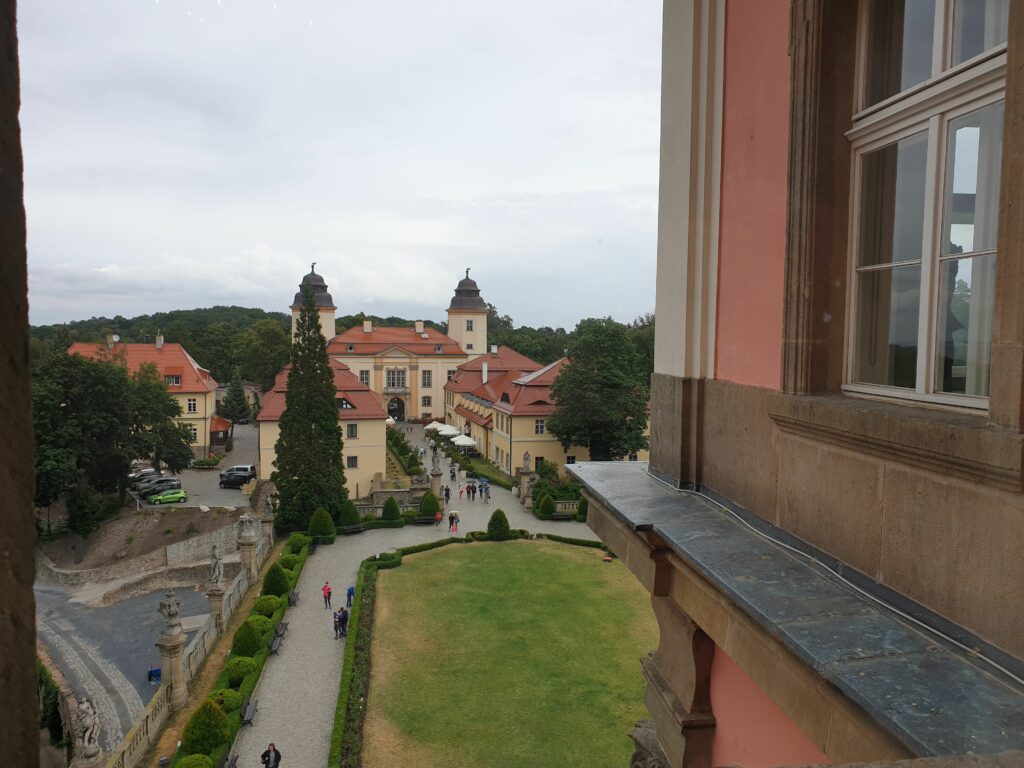 Widok z wysokiego okna Zamku Książ na rozległy dziedziniec główny, skomplikowaną architekturę dachów, bocznych skrzydeł i bramy. W tle zalesione wzgórza Książańskiego Parku Krajobrazowego pod chmurnym niebem. Kontrast dla surowych podziemi.