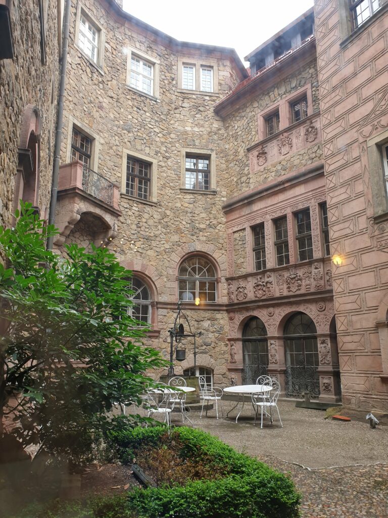 Kameralny dziedziniec wewnętrzny Zamku Książ, otoczony surowymi kamiennymi murami i bogato zdobioną elewacją z rzeźbionymi detalami. W centrum znajduje się stara studnia oraz kute, białe meble ogrodowe.