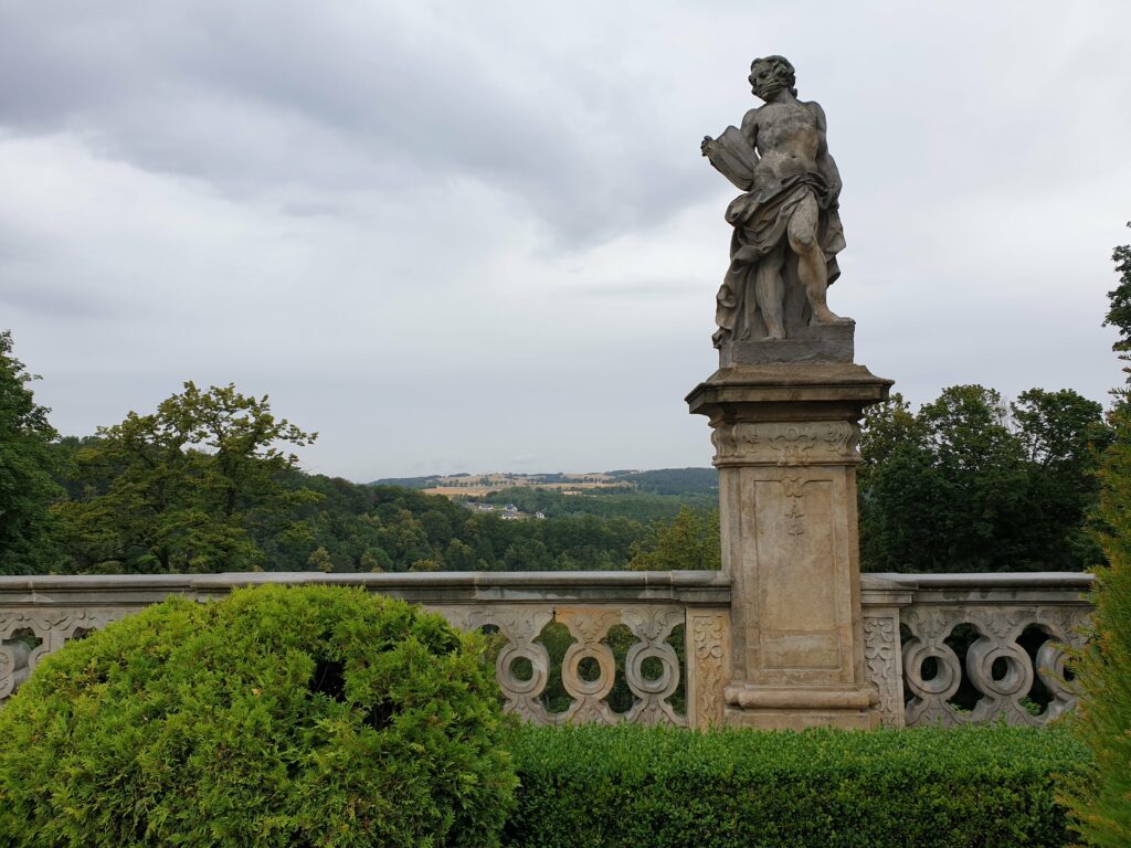 Zabytkowa, barokowa rzeźba na zdobionej balustradzie tarasu Zamku Książ, spoglądająca na zalesioną dolinę pod zamkiem. Ujęcie ze ścieżki prowadzącej do punktu widokowego na skale Olbrzyma.