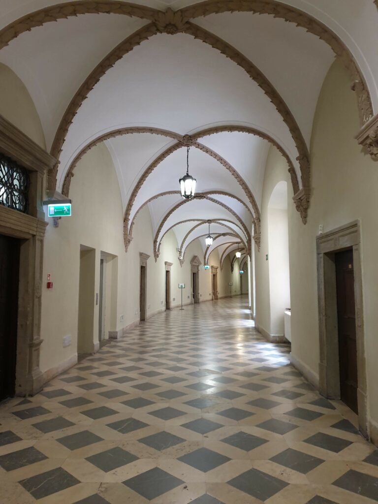 Długi, jasny korytarz na trasie zwiedzania wewnątrz Zamku Książ. Wyróżnia się ozdobnym sklepieniem, stylowymi latarniami i posadzką ułożoną w geometryczną szachownicę.