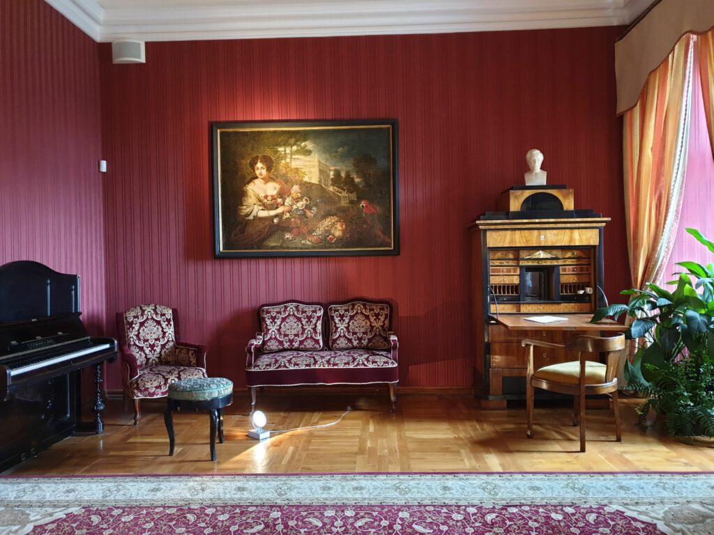 Opulentne wnętrze Zamku Książ, prawdopodobnie Salon Daisy, z pociągniętym czerwoną tapetą i zdobionym sufitem. Widać zabytkowe pianino, orzechowy sekretarzyk, fotele z tapicerką we wzory oraz centralny portret księżnej Daisy von Pless.