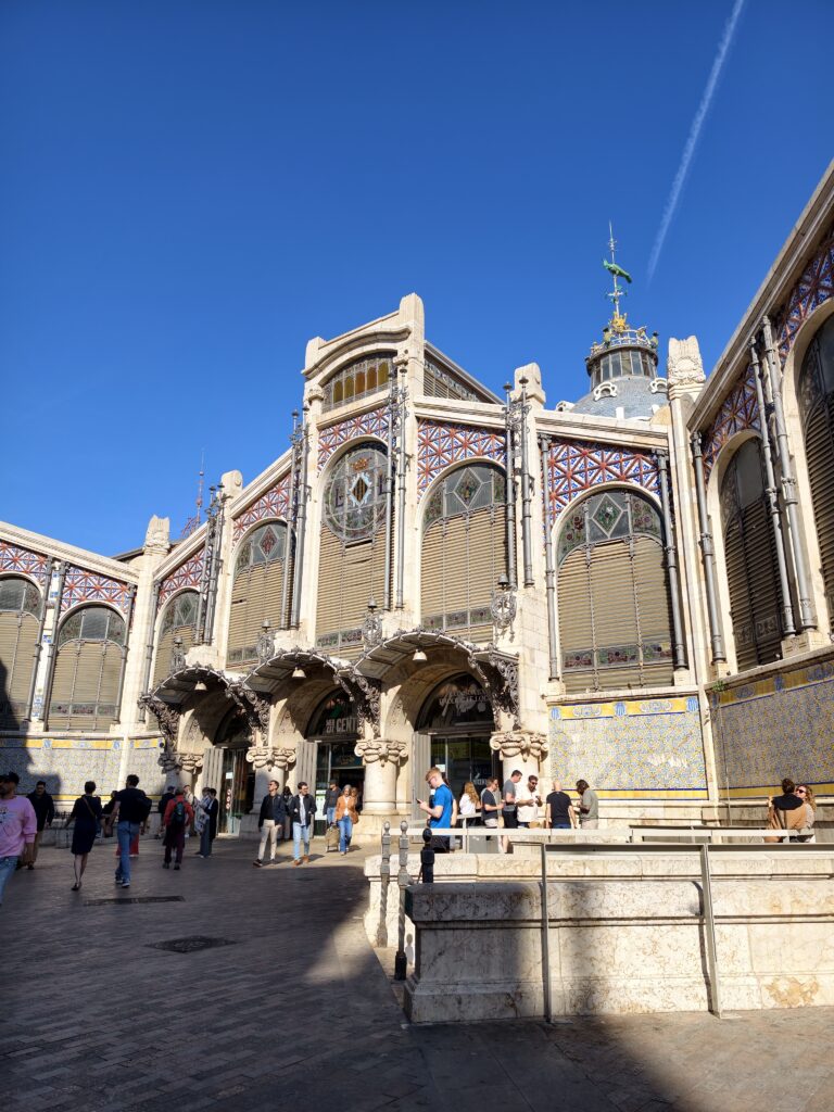 Modernistyczna fasada głównego wejścia do hali targowej Mercado Central w Walencji, wyróżniająca się monumentalną konstrukcją ze szkła, żelaza i zdobieniami z ceramicznych płytek.