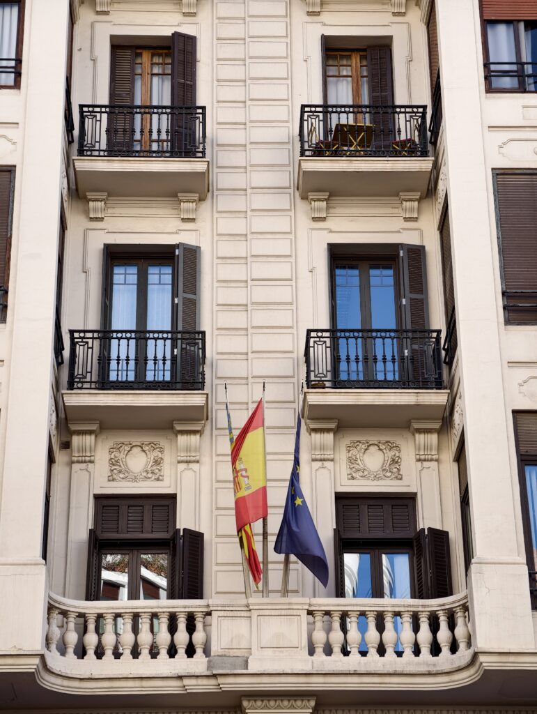 Jasna, elegancka fasada historycznej kamienicy w Walencji, na której wyróżniają się tradycyjne, kute z żelaza balkony i wysokie okna z okiennicami.