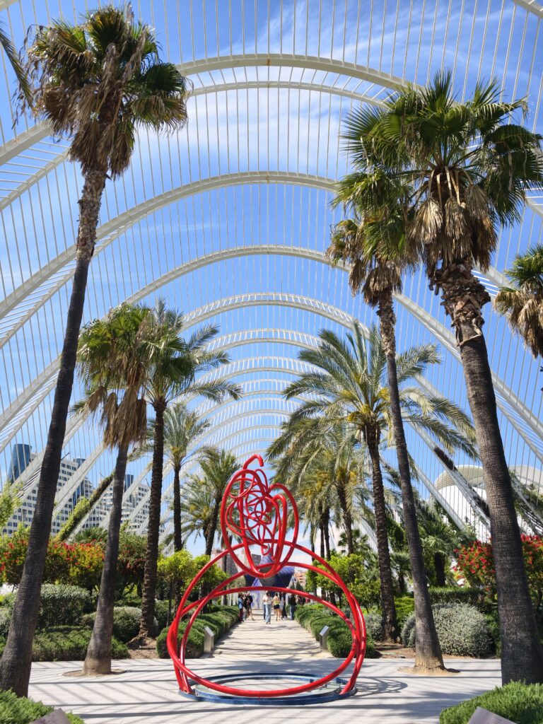 Długa aleja spacerowa pod białymi, ażurowymi łukami konstrukcji El Umbracle w Walencji, otoczona rzędami zielonych palm i gęstą, śródziemnomorską roślinnością.