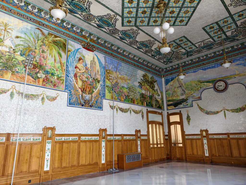 Wnętrze głównego holu zabytkowego dworca Estación del Norte w Walencji z widocznymi bogatymi, modernistycznymi zdobieniami z drewna oraz kolorowymi mozaikami ceramicznymi.