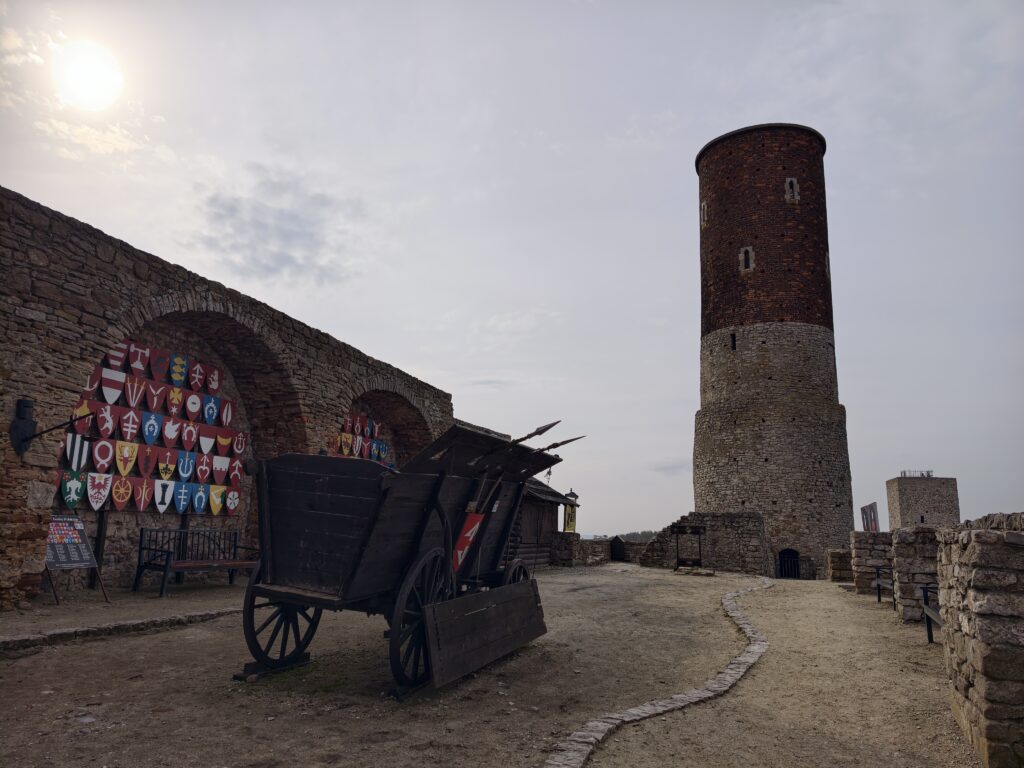 Drewniany wóz, kolorowe tarcze herbowe i wysoka ceglana wieża na dziedzińcu warowni w Chęcinach