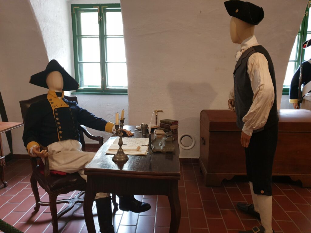 Manekiny w historycznych mundurach przy stole z dokumentami na wystawie w Twierdzy Kłodzkiej.