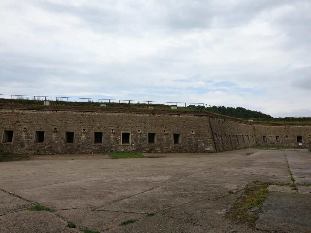 Długi budynek kazamat na dziedzińcu Twierdzy Kłodzkiej, z wieloma małymi oknami.