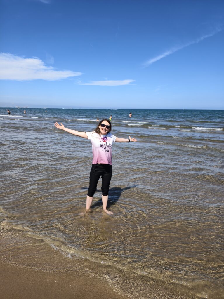 Kobieta spacerująca brzegiem morza i mocząca bose stopy w płytkich falach na szerokiej plaży Las Arenas w Walencji.