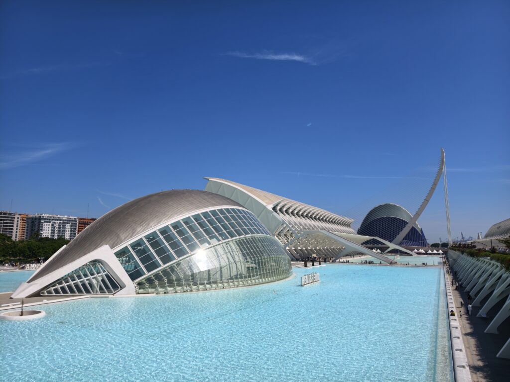 Futurystyczny budynek L’Hemisfèric w Walencji w kształcie na wpół otwartego oka, odbijający się w otaczającym go płytkim, lazurowym basenie wodnym w słoneczny dzień.