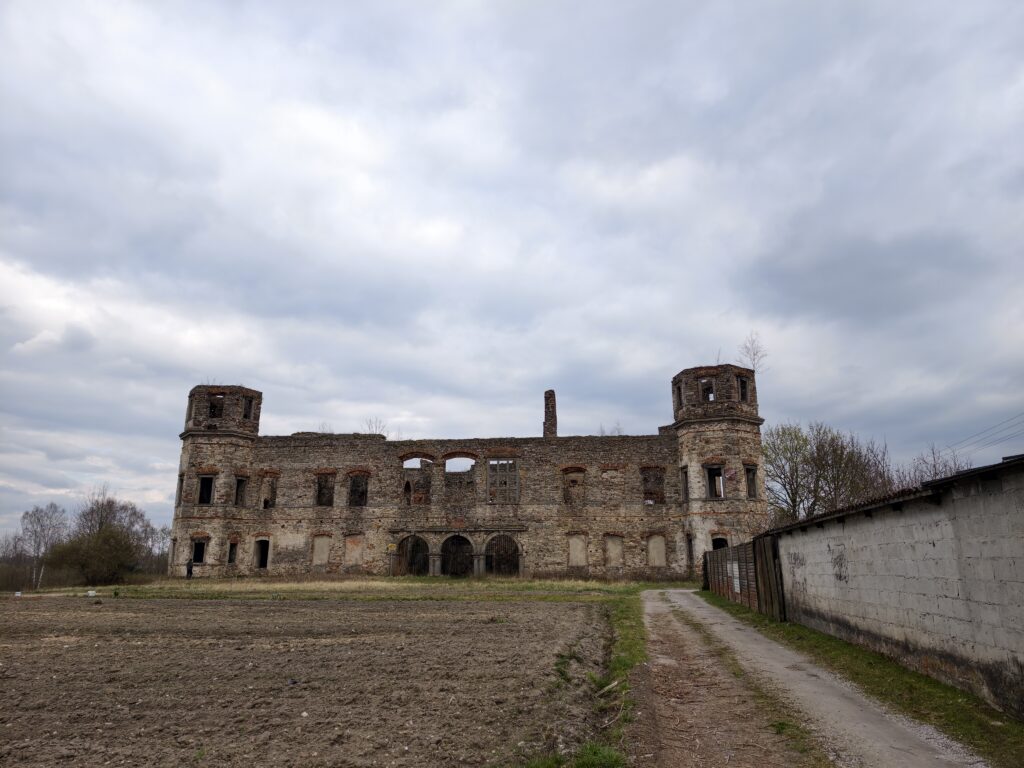 Majestatyczna, ogołocona z dachu fasada i dwie sześcioboczne wieże narożne ruin Pałacu Tarłów w Podzamczu Piekoszowskim, świętokrzyskie, na tle wiosennej zieleni i zachmurzonego nieba.