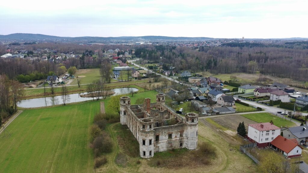 Szersze ujęcie z drona przedstawiające ruiny Pałacu Tarłów w Podzamczu Piekoszowskim na tle wsi, pól i pobliskiego stawu.