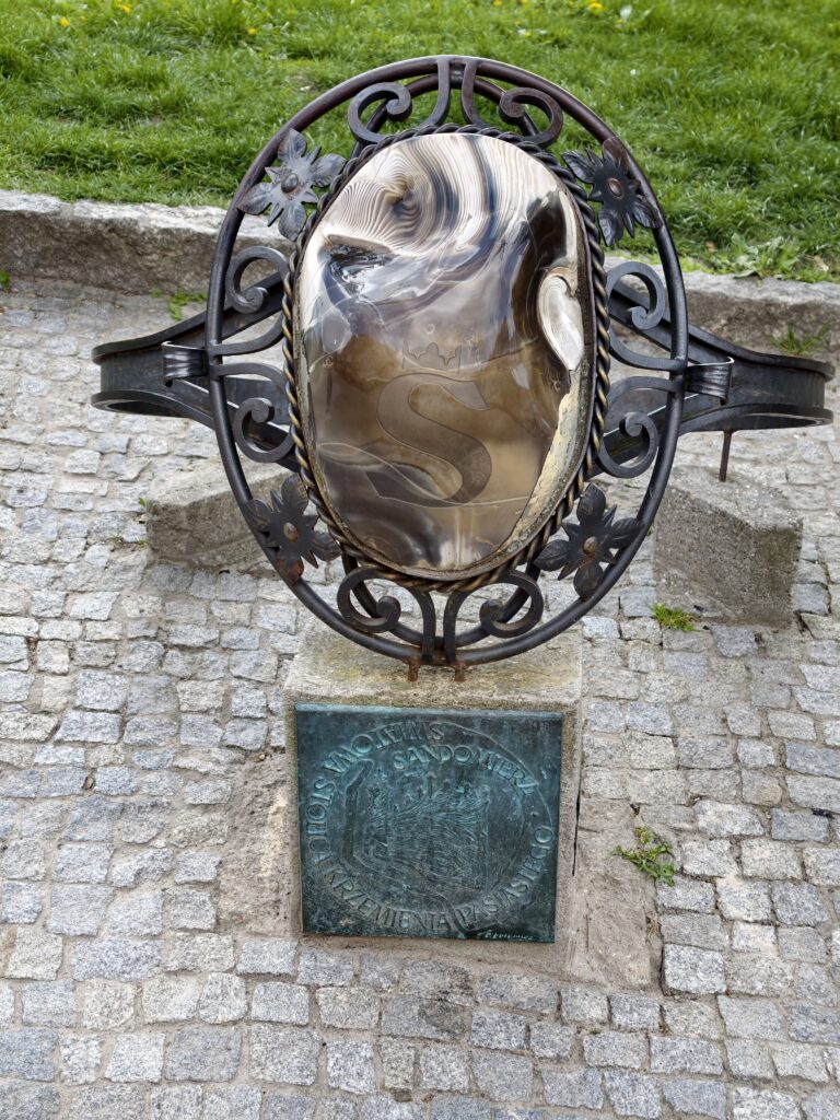 Duża, metalowa rzeźba od frontu w kształcie pierścienia z krzemieniem pasiastym na skwerze w Sandomierzu.