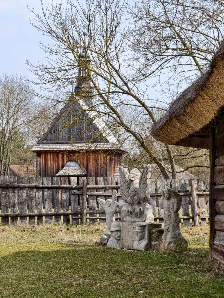 Zabytkowe rzeźby kamienne na pierwszym planie na tle drewnianego kościoła z Rogowa w Muzeum Wsi Kieleckiej