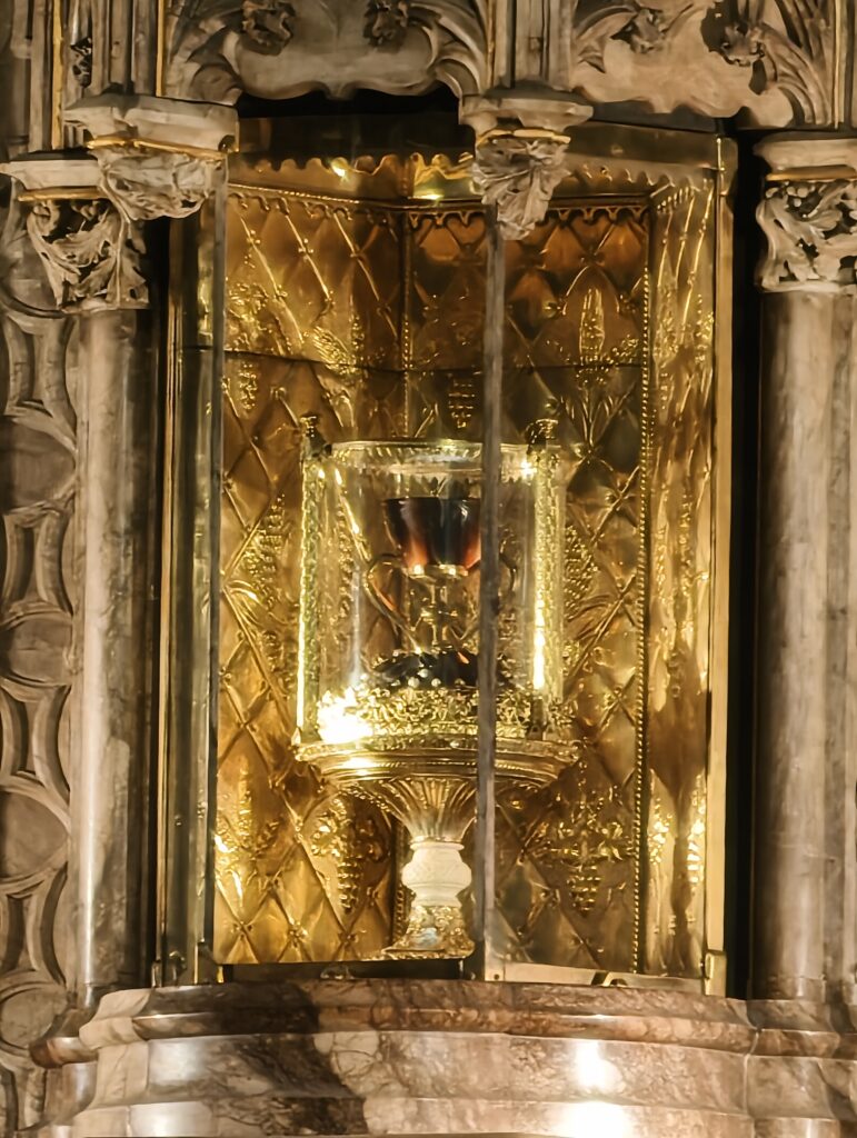 Zbliżenie na zabytkowy kielich wykonany z ciemnego agatu, osadzony na złotej podstawie bogato zdobionej perłami i kamieniami szlachetnymi, oświetlony wewnątrz gabloty.