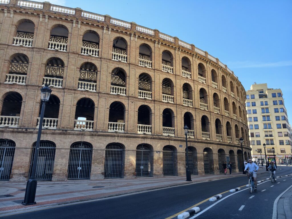 Monumentalna, okrągła fasada areny Plaza de Toros w Walencji, zbudowana z czerwonej cegły z rzędami klasycznych łuków, przypominająca rzymskie Koloseum.
