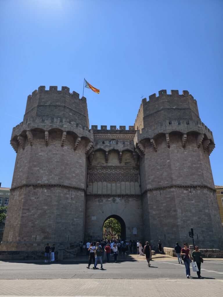 Potężne, średniowieczne wieże Torres de Serranos w Walencji, zbudowane z jasnego kamienia, z widocznymi gotyckimi łukami i blankami na szczycie.