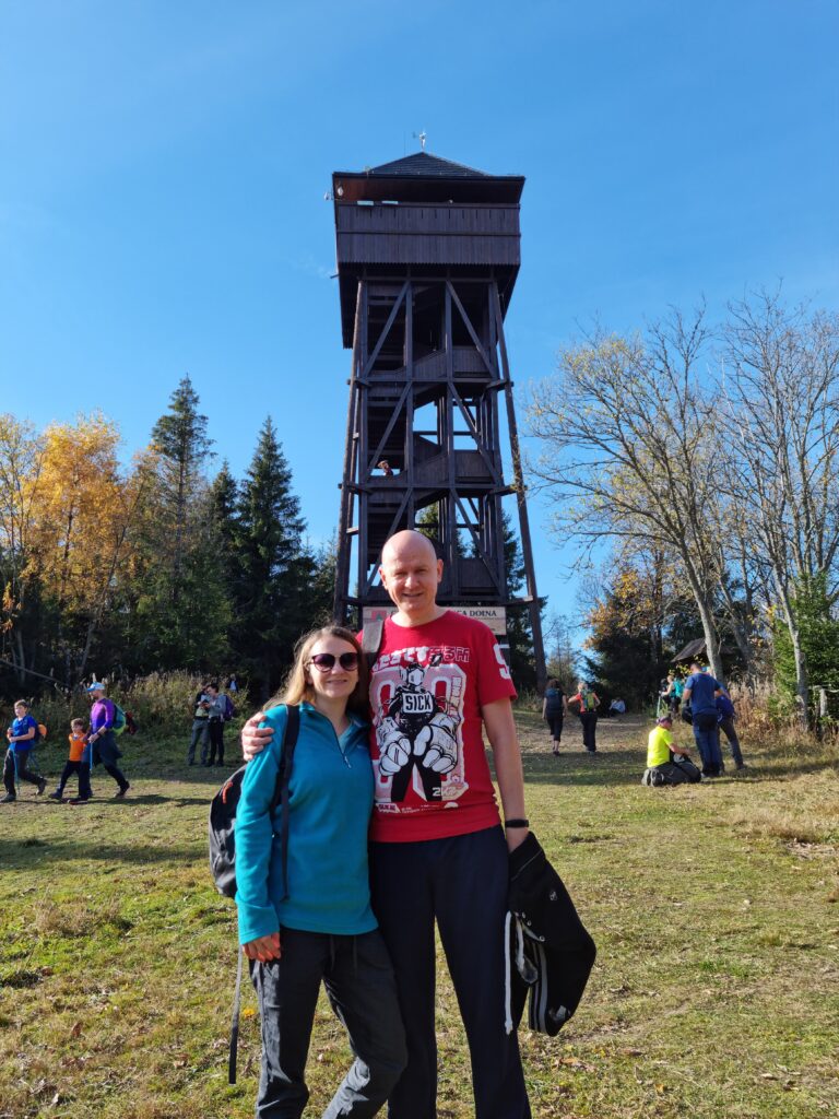 Uśmiechnięta para turystów stojąca na trawiastej polanie przed wysoką, drewnianą wieżę widokową na szczycie Magurek w Gorcach.