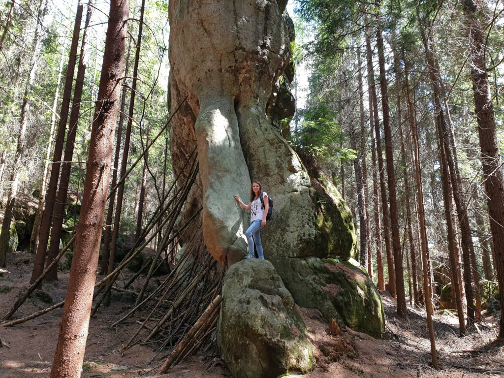 Uśmiechnięta kobieta w stroju trekkingowym i z plecakiem pozuje na ścieżce obok potężnego skalnego borowika.