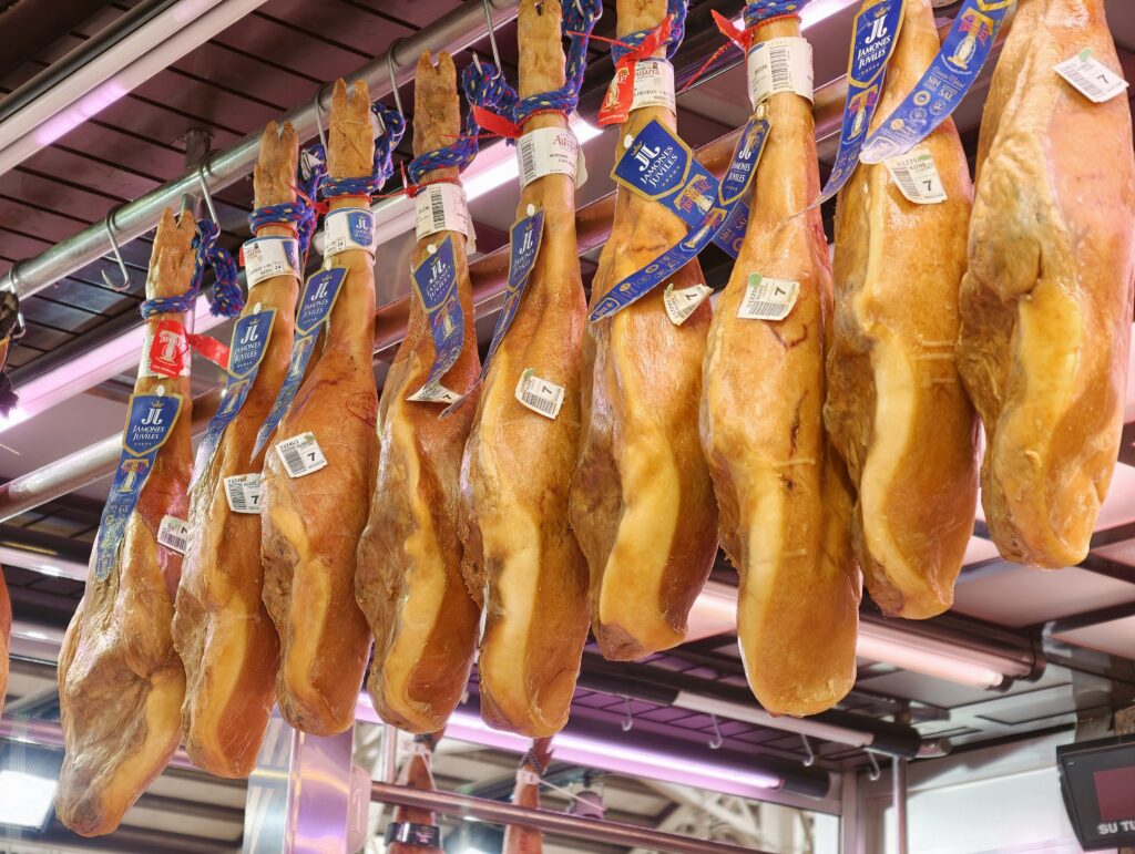 Tradycyjne hiszpańskie dojrzewające szynki jamón ibérico, zawieszone na hakach pod sufitem na jednym ze stoisk mięsnych w hali targowej Mercado Central w Walencji.