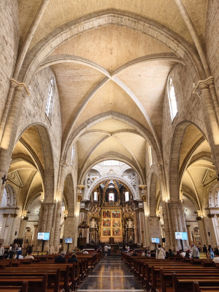 Szeroki kadr przedstawiający przestronne, gotyckie wnętrze Katedry w Walencji z rzędem masywnych, kamiennych kolumn, wysokim sklepieniem żebrowym i światłem wpadającym przez witraże.
