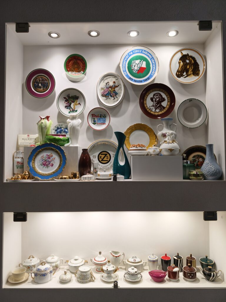 Szklana gablota z pamiątkowymi talerzami, wazonami i okolicznościową porcelaną na wystawie w muzeum w Ćmielowie.