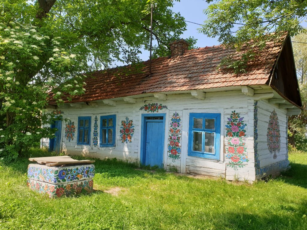 Stara, drewniana malowana chata w Zalipiu z błękitnymi okiennicami i kwiecistymi wzorami oraz ozdobioną studnią na trawniku.