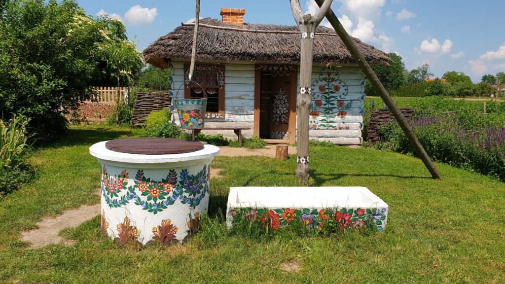 Ręcznie malowana studnia z kwiatami i drewniana chata pod strzechą w zagrodzie Felicji Curyłowej w Zalipiu.