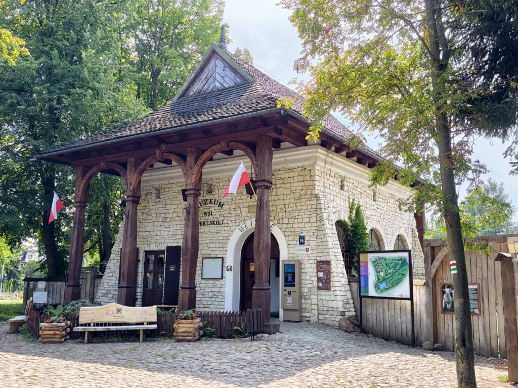 Kamienny budynek wejściowy do Muzeum Wsi Lubelskiej w Lublinie z drewnianym podcieniem, kasą biletową i tablicą informacyjną