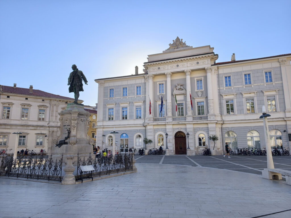 Przestronny Plac Tartiniego w Piranie z brązowym pomnikiem kompozytora i reprezentacyjnym budynkiem ratusza w tle.