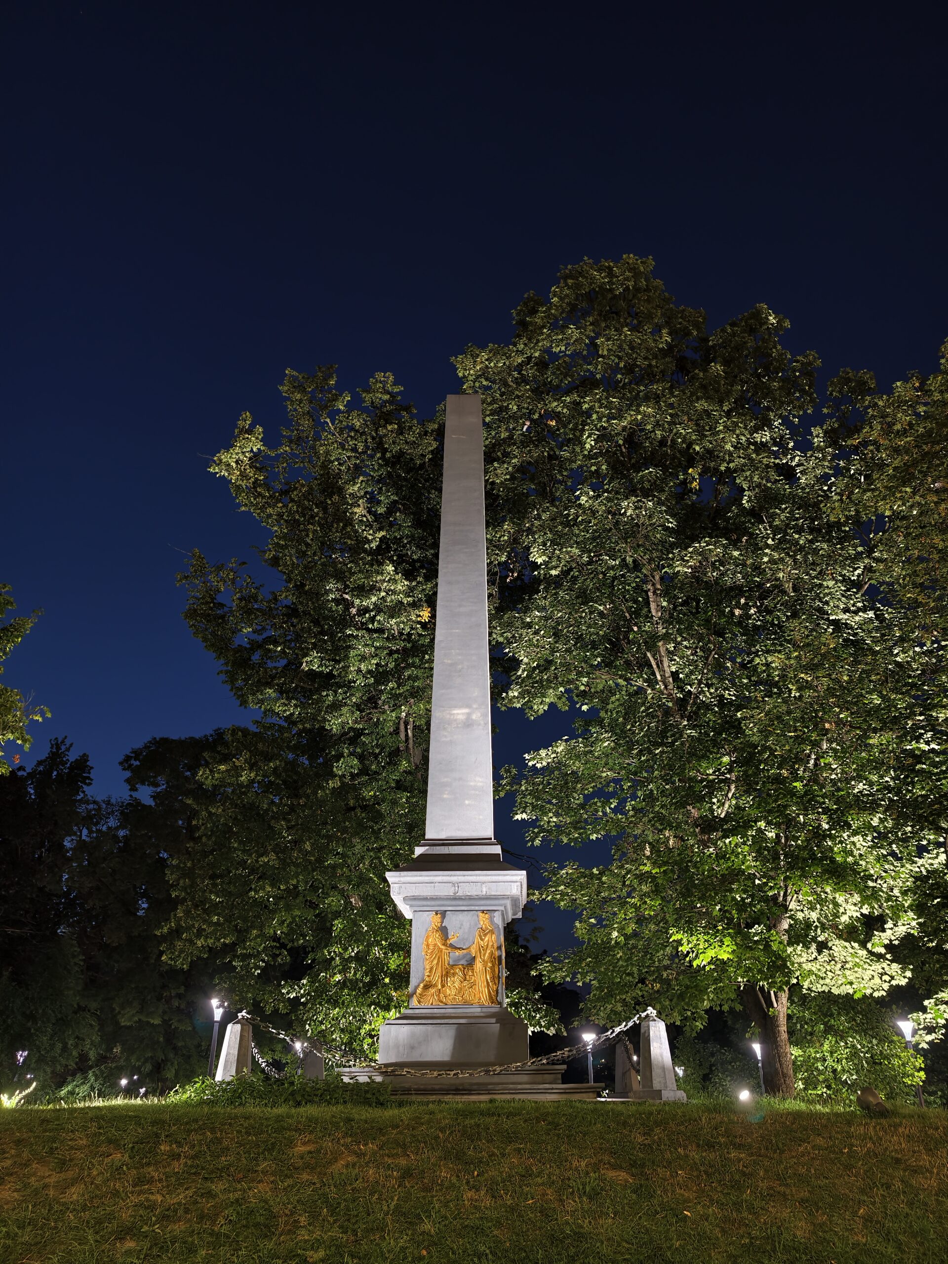 Oświetlony nocą obelisk pomnika Unii Lubelskiej na Placu Litewskim w Lublinie, otoczony drzewami