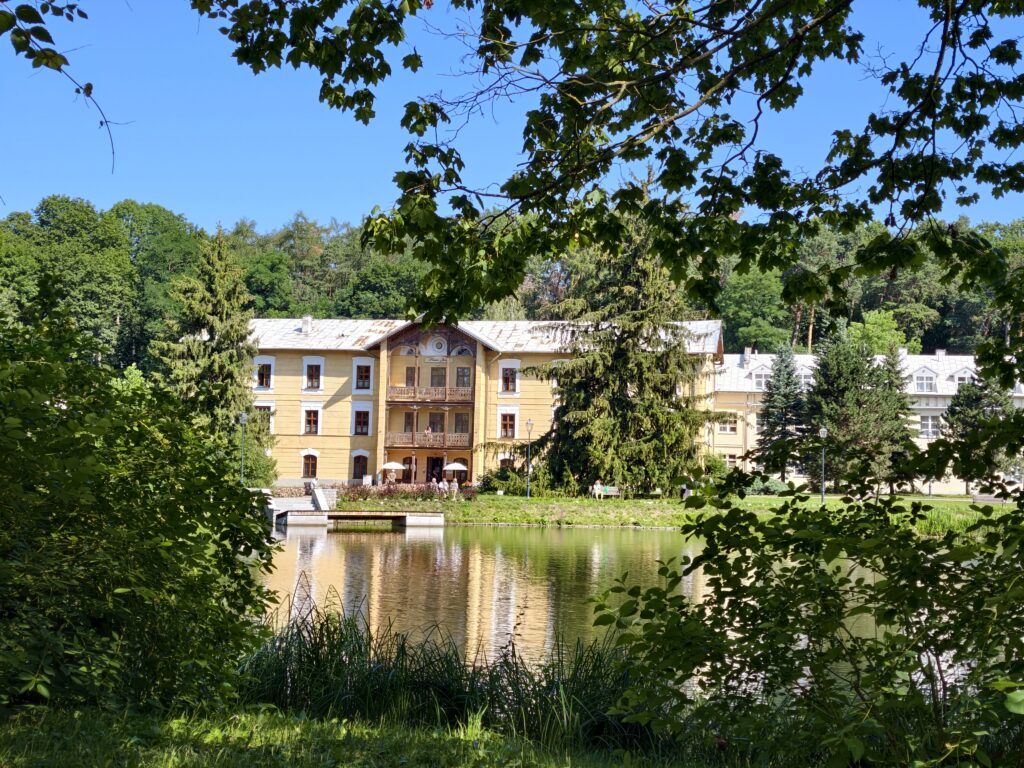 Sanatorium Książę Józef w Nałęczowie widoczne znad brzegu stawu przez naturalną ramę z zielonych liści drzew.
