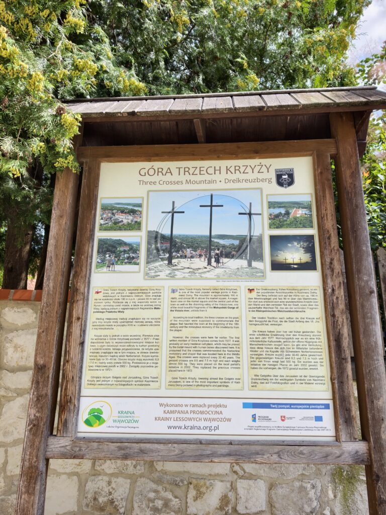 Drewniana tablica edukacyjna z napisem Góra Trzech Krzyży. Na tablicy widnieją historyczne teksty w trzech językach oraz zdjęcia archiwalne i współczesne z widokiem na krzyże i panoramę.