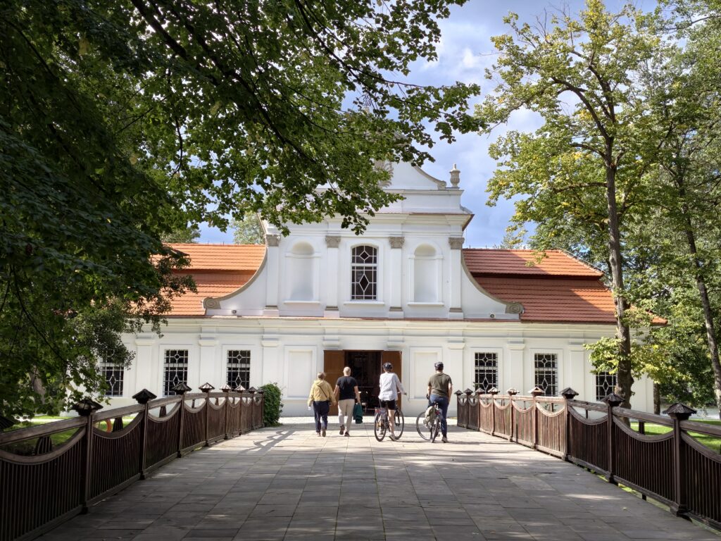 Turyści z rowerami idący drewnianym mostem w kierunku Kościoła na Wodzie w Zwierzyńcu