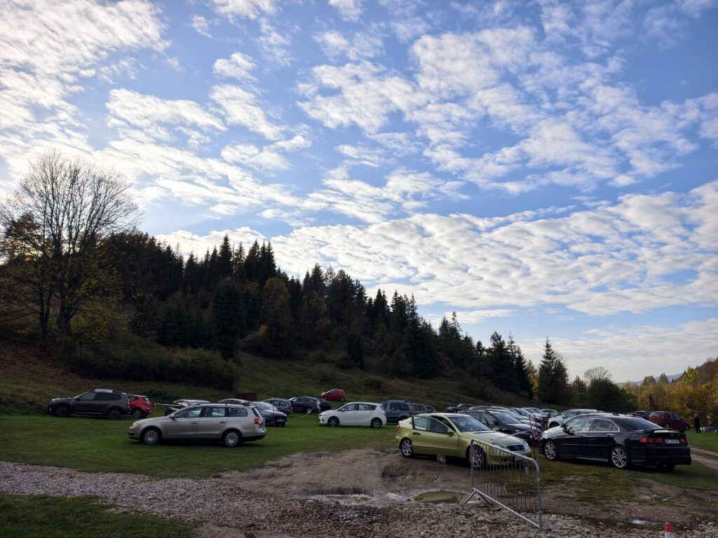 Samochody na trawiastym parkingu przy Wąwozie Homole w Jaworkach, za nimi zalesione wzgórze