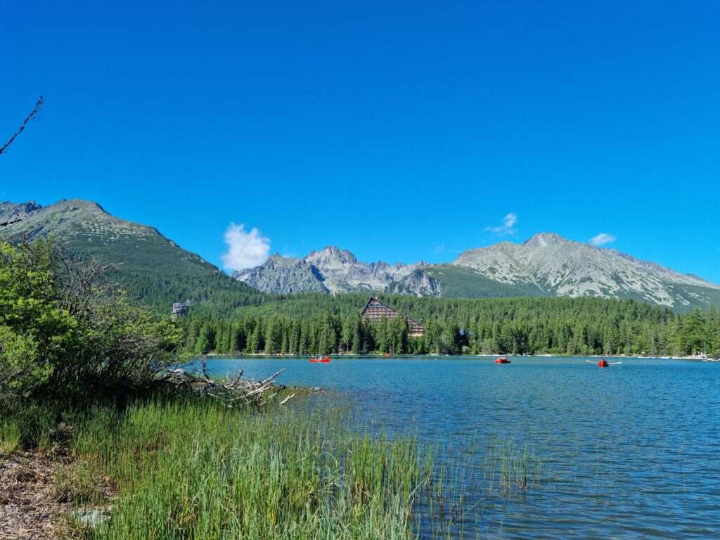 Szczyrbskie Jezioro latem: porośnięty trawą brzeg, czerwone łódki na wodzie i Tatry Wysokie w tle