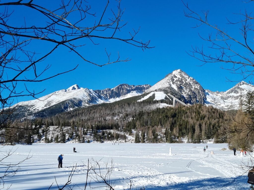 Zamarznięte i przysypane śniegiem Szczyrbskie Jezioro z ludźmi i bałwanami na lodzie, w tle ośnieżone Tatry i skocznie